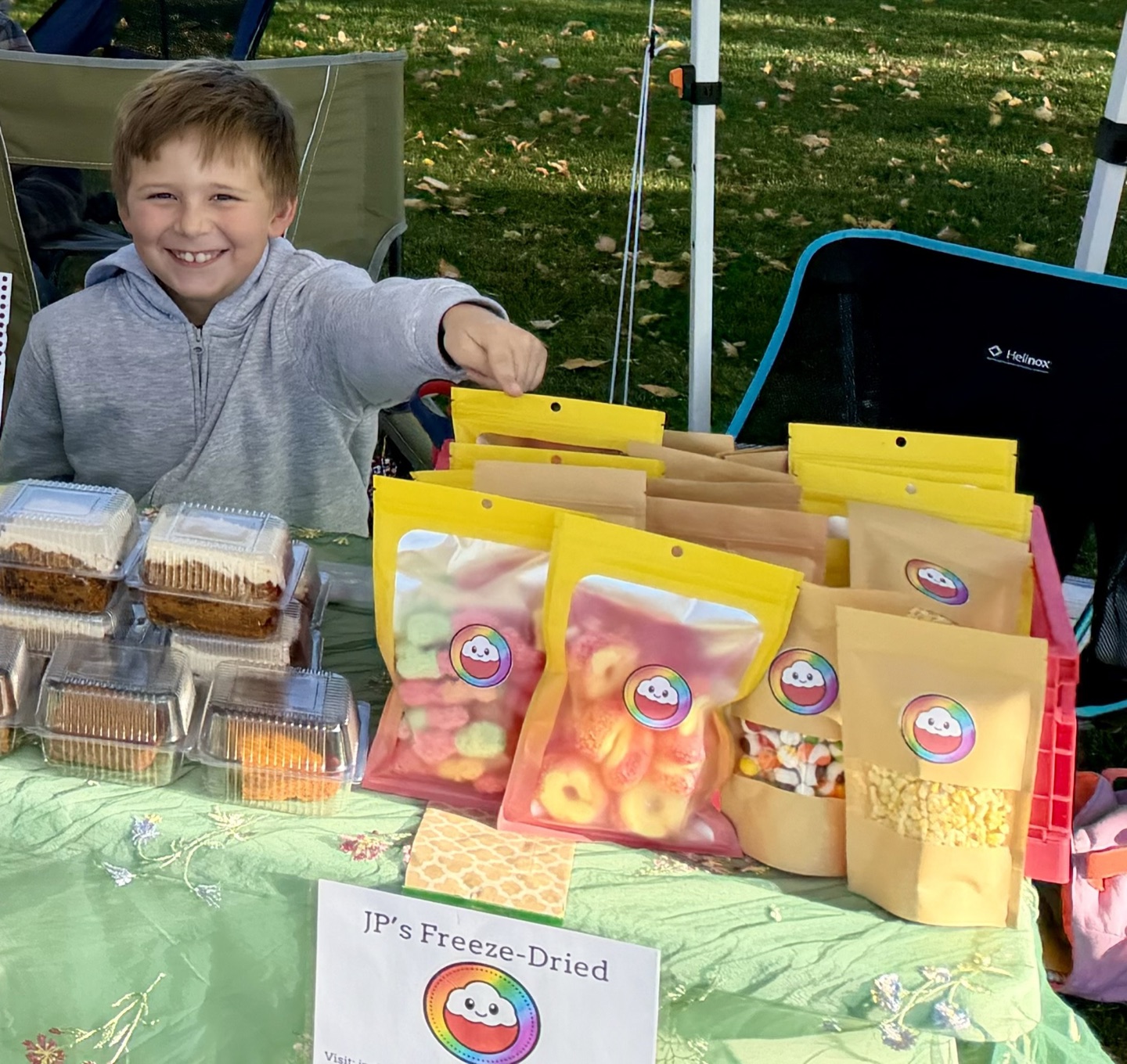 JP grinning and pointing at the camera behind his market table, filled with yellow JP's Freeze-Dried bags, including a bag of freeze-dried corn